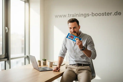 Mann isst Proteinriegel am Schreibtisch mit Laptop, ideal für Sportnahrung und Energiebooster