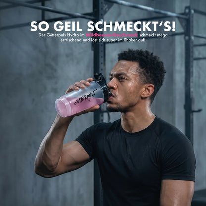 Muskulöser Mann trinkt pinken Göttelpuls Hydro Wildbeeren-Booster im Fitnessstudio